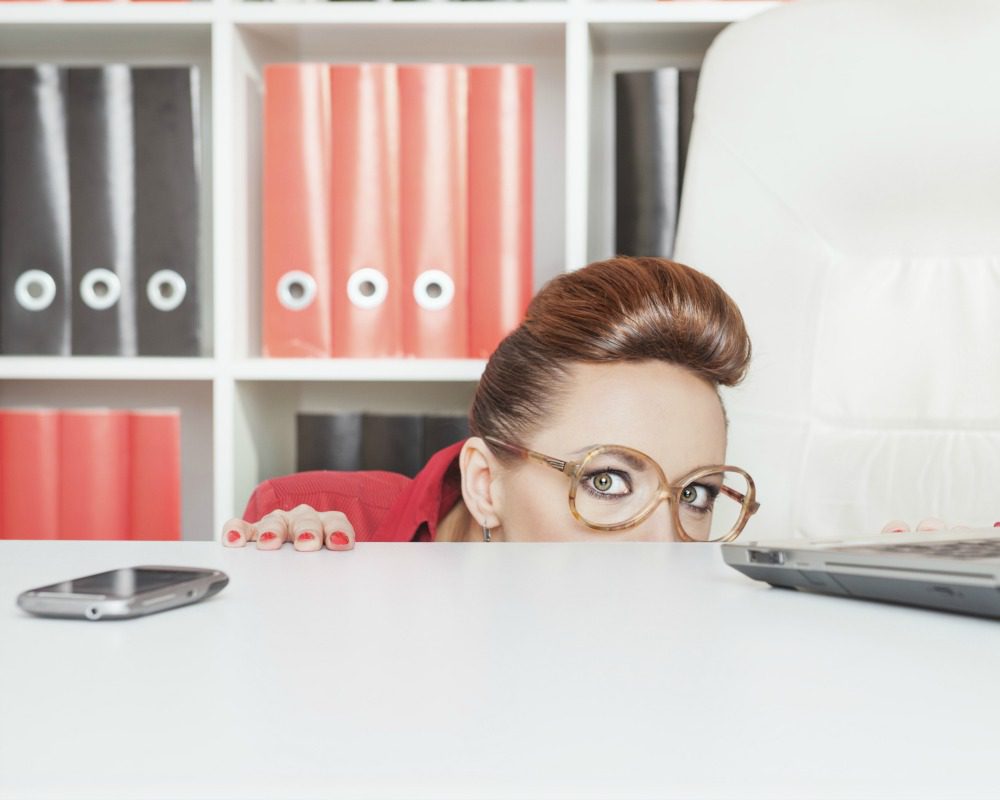 Business woman with glasses hiding behind table and afraid