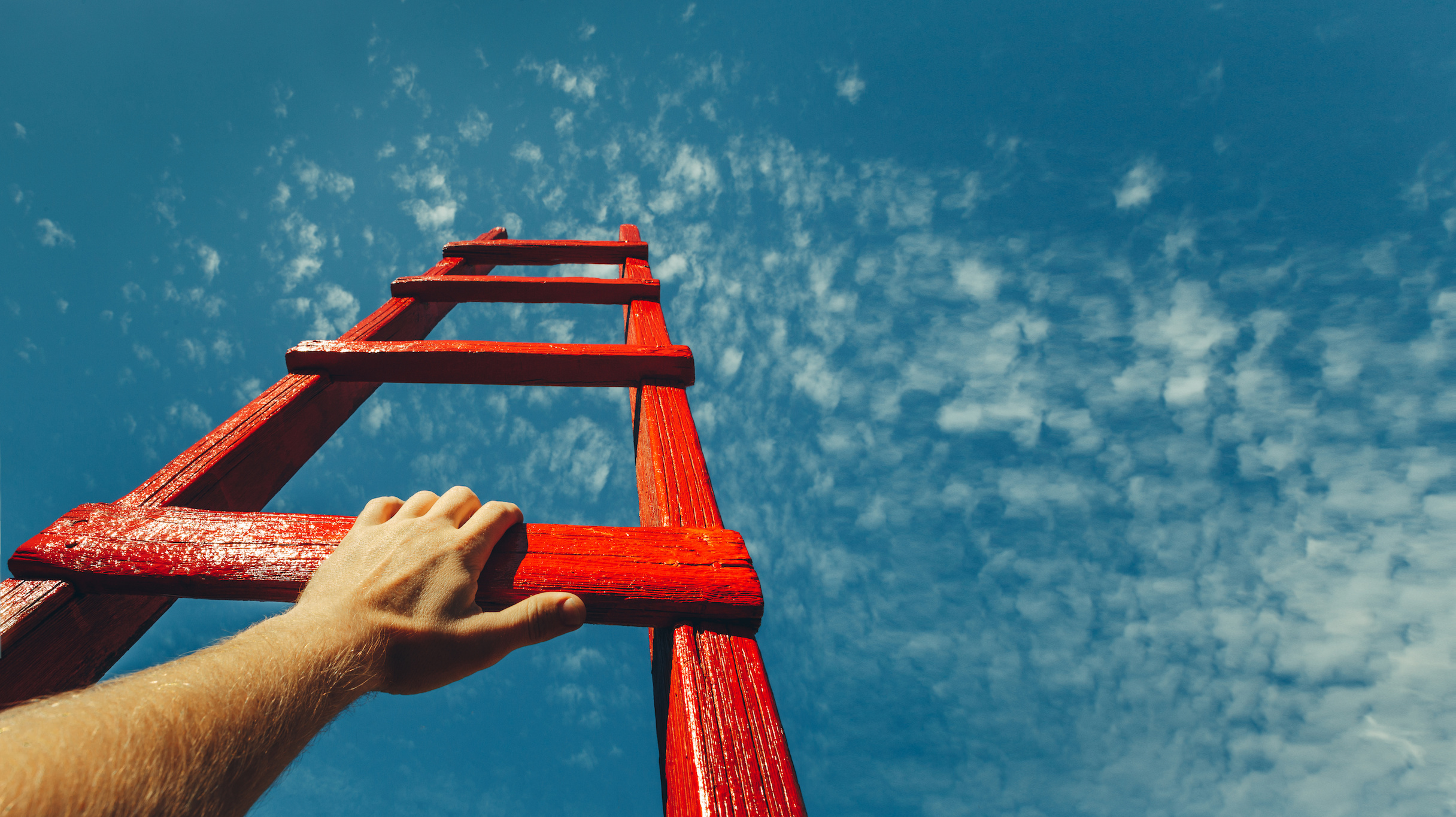 A hand holds onto the crossbar of a red wooden ladder leading to the blue sky